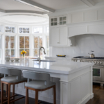 Bright kitchen island with sink; chairs, range, bay window.